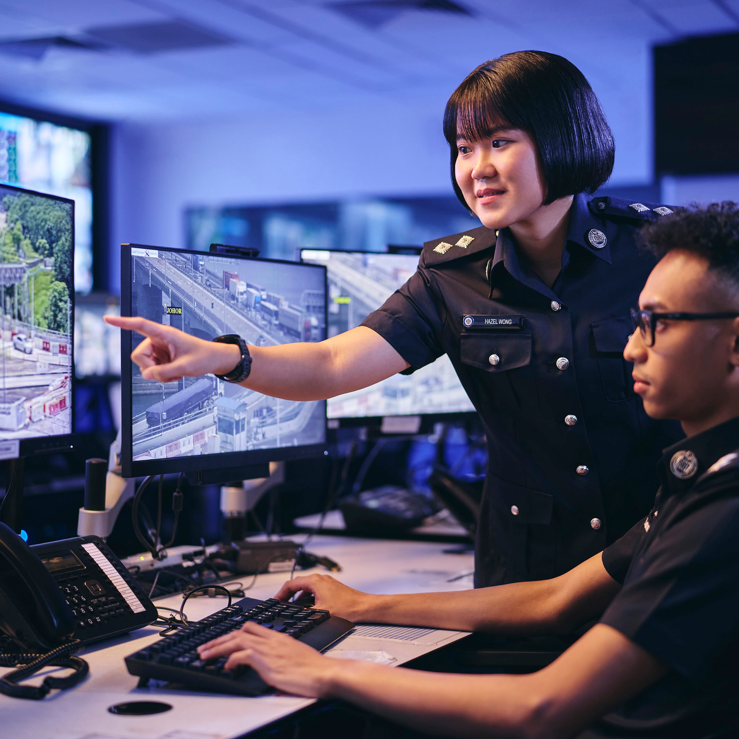 Two officers in dark uniforms in a control room, one pointing at a monitor, one at a keyboard.